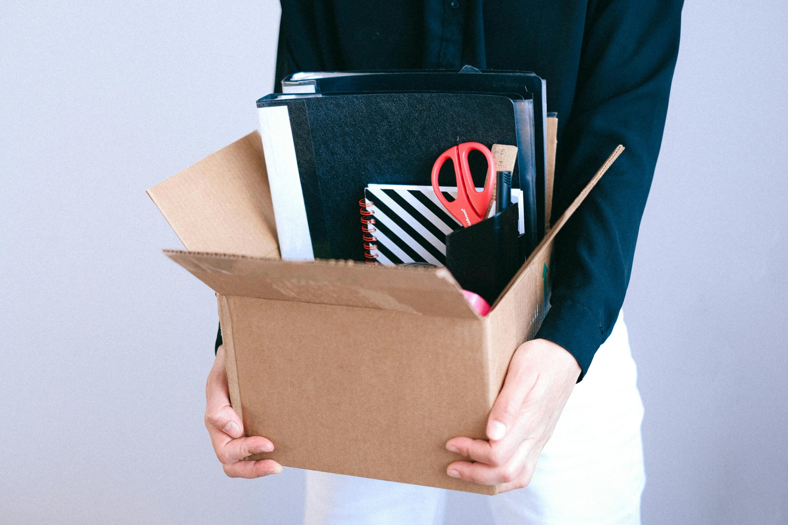 A person carrying a cardboard box filled with office supplies and notebooks after being laid off from work.
