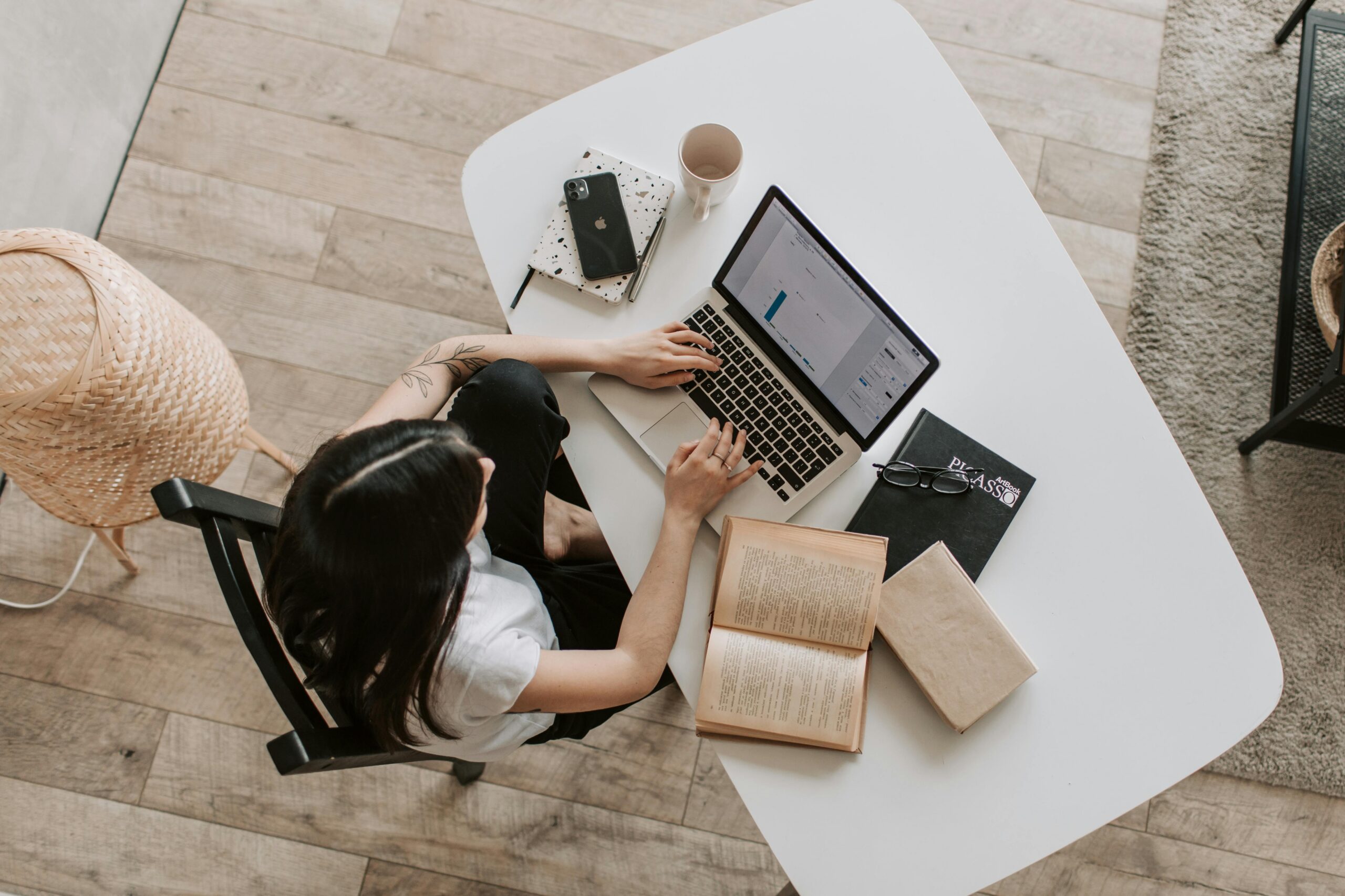Overhead view of a woman working alone on a laptop at a white desk surrounded by open books, a phone, and a notebook.